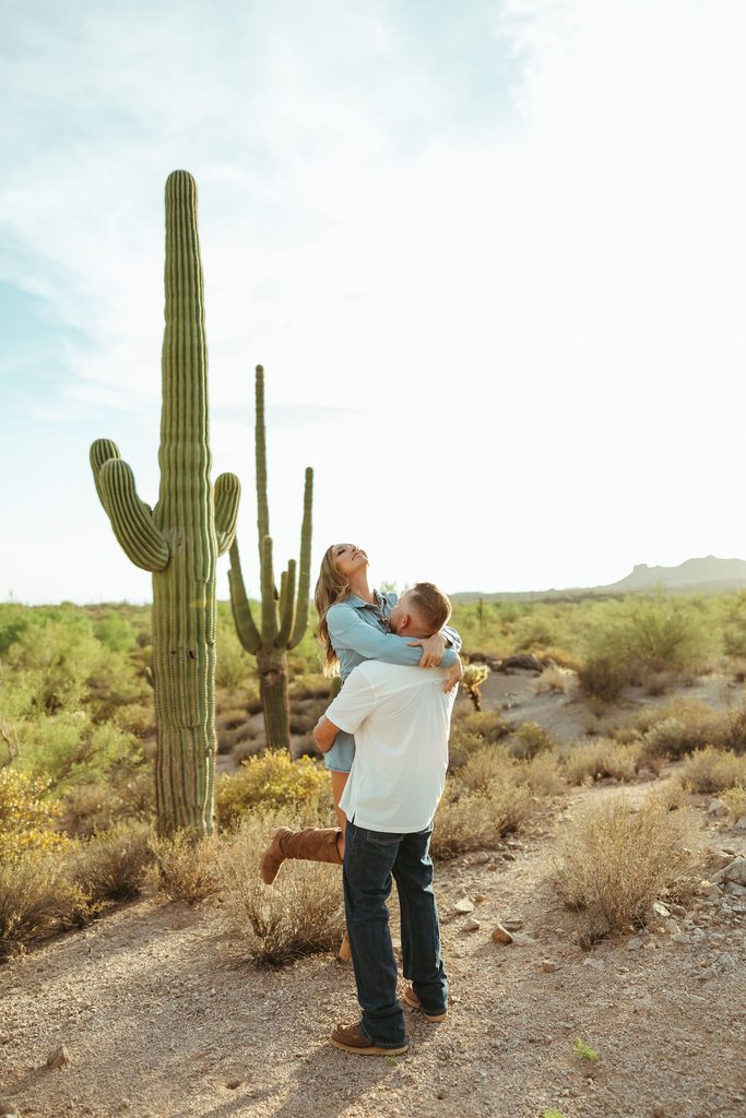 Superstition Mountains couples session featuring a playful moment as the bride-to-be is lifted by her fiancé in the Arizona desert surrounded by tall saguaros.