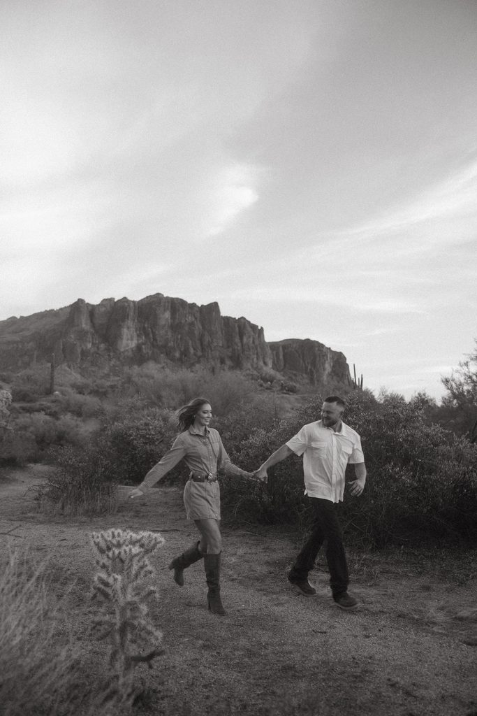 Arizona couples photographer capturing a romantic black and white moment as a couple walks hand in hand through the desert beneath the Superstition Mountains.