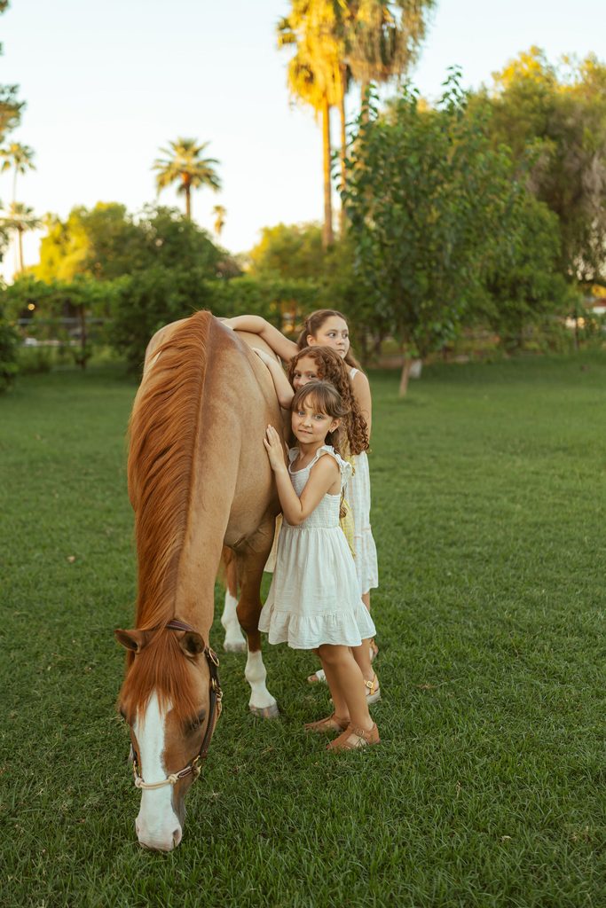 Gilbert family photographer capturing sisters standing beside their horse during a golden hour family photography session at Floyd Family Farms.